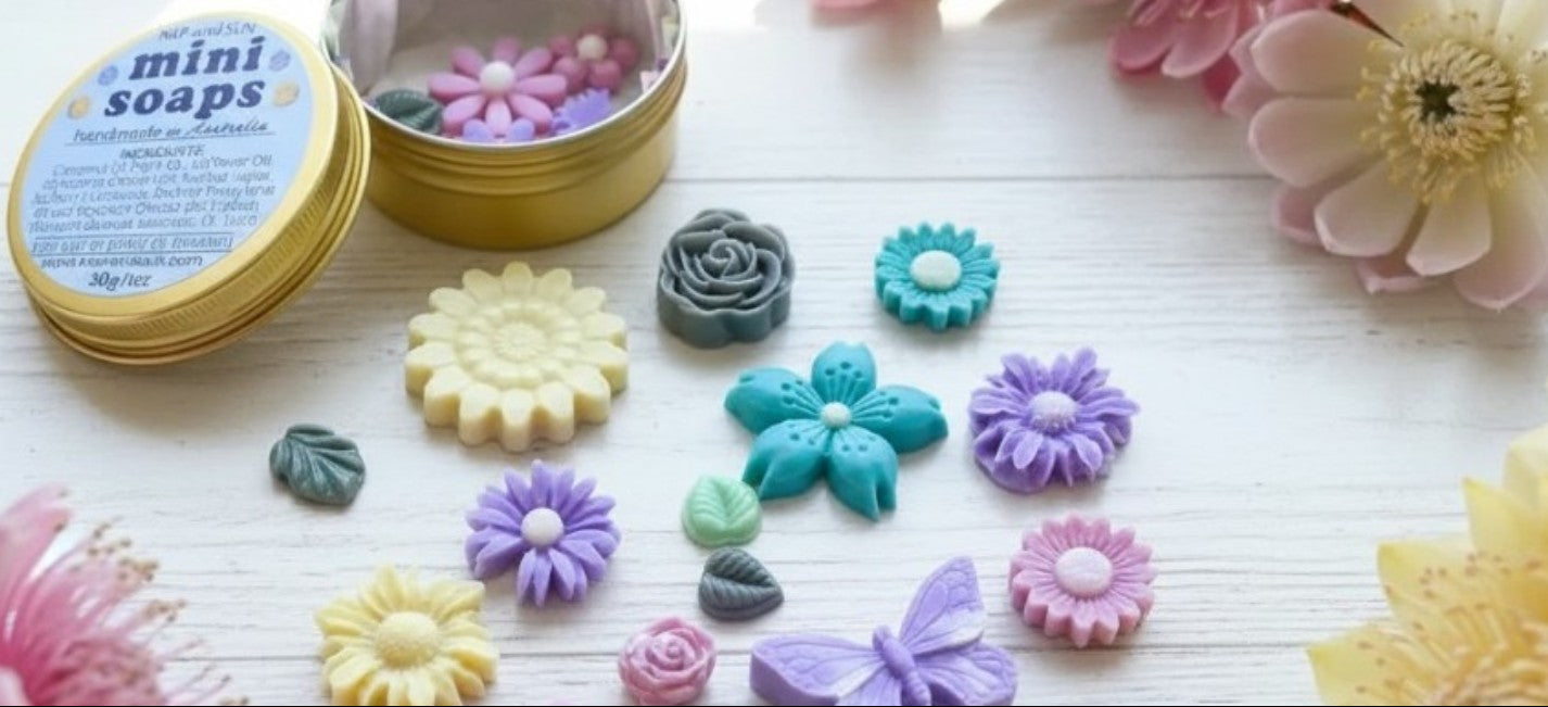  A set of colorful mini flower-shaped soaps displayed on a white wooden surface next to their gold tins, surrounded by decorative pink and yellow flowers.
