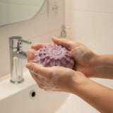A person's hands lathered with white soap foam holding a textured, lavender-purple hand soap bar in a bathroom setting with a sink and faucet in the background.