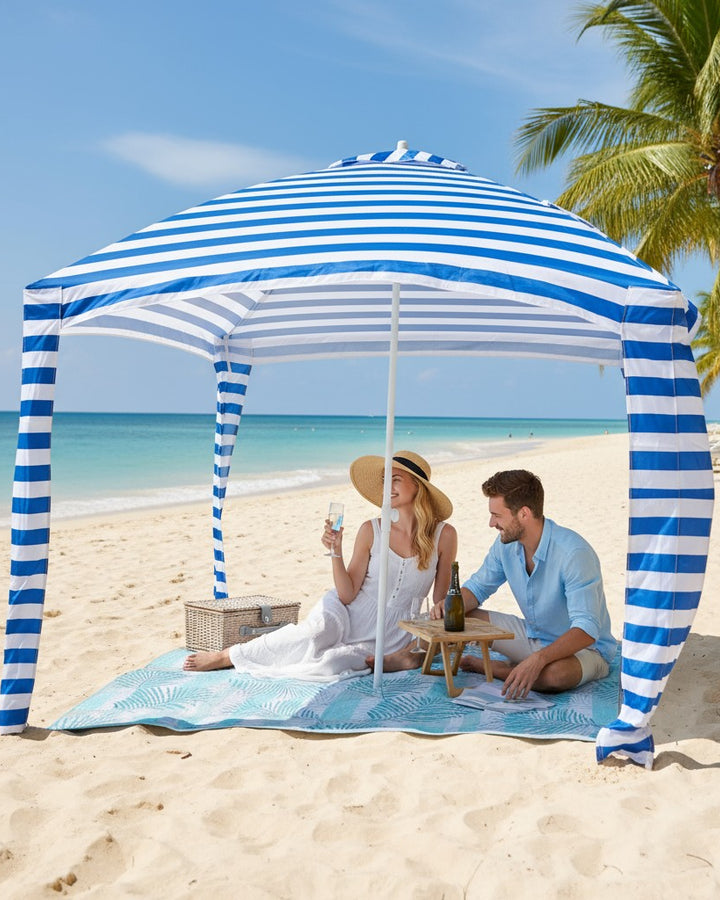 Blue and white striped beach canopy with a couple sitting underneath on a picnic blanket on a sandy beach.