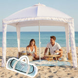 Leafy picnic blanket set up on a sandy beach under a cabana, with people relaxing and enjoying a picnic.