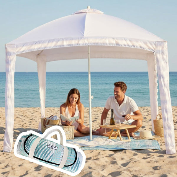Leafy picnic blanket set up on a sandy beach under a cabana, with people relaxing and enjoying a picnic.