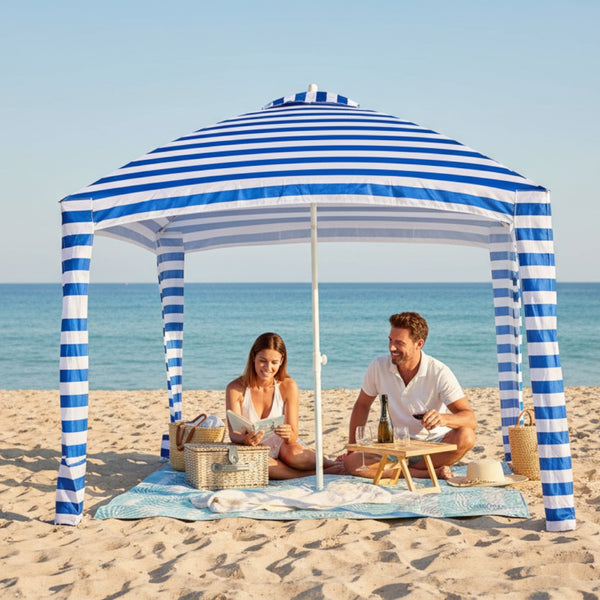 Leafy picnic blanket set up on a sandy beach under a cabana, with people relaxing and enjoying a picnic.