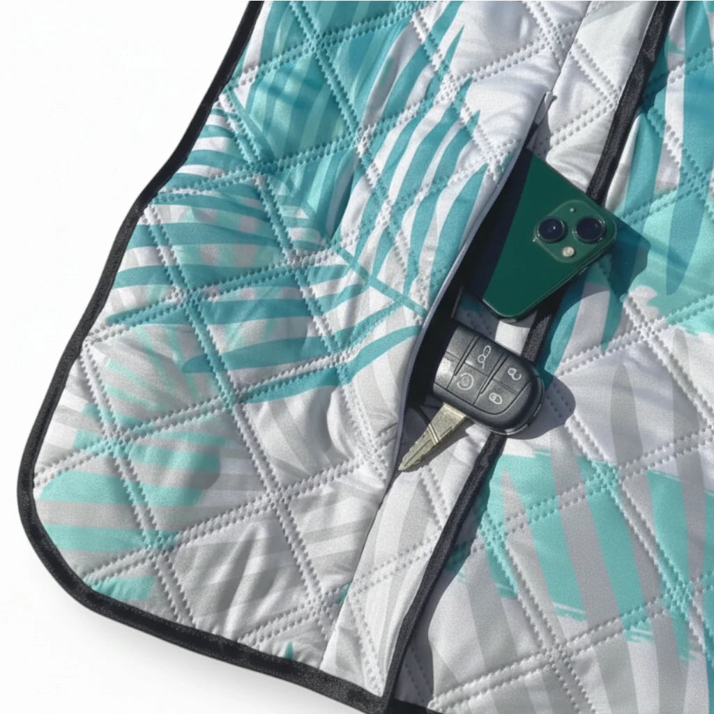 Close-up of the zip pocket on a light green palm leaf-patterned picnic blanket, holding a phone and car key.