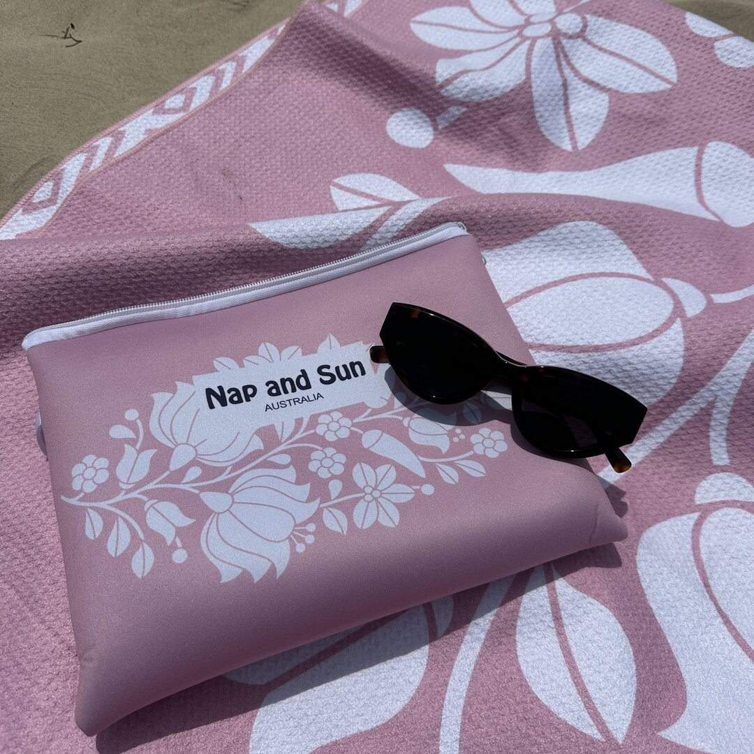 close up of Nap and Sun Pink Bloom sand-free beach towel. bright pink colour towel with white flower pattern. Matching neoprene bag is lying on a towel with a sunglasses on top