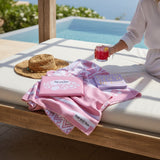 woman holding a drink on a sunbed with nap and sun brand pink towel and book by a poolside.
