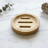 A round natural bamboo soap dish with three horizontal slots, placed on a white marble surface with blurred decorative eucalyptus leaves and a towel in the background.