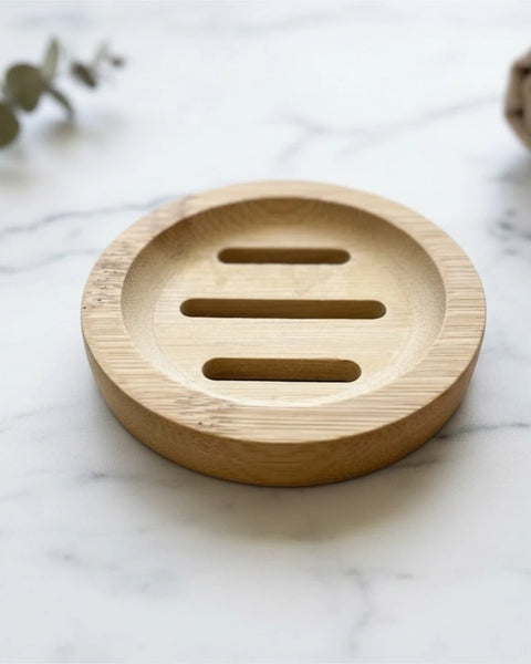 A round natural bamboo soap dish with three horizontal slots, placed on a white marble surface with blurred decorative eucalyptus leaves and a towel in the background.