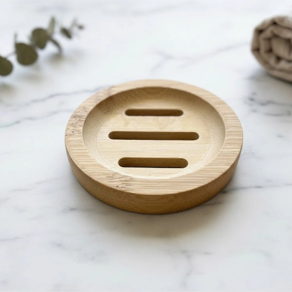 A round natural bamboo soap dish with three horizontal slots, placed on a white marble surface with blurred decorative eucalyptus leaves and a towel in the background.