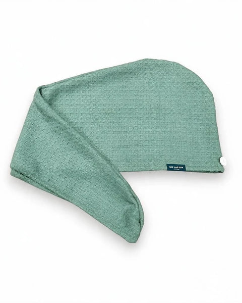 A sage green waffle hair towel wrap with a small dark blue brand tag, shown on a clean white background.