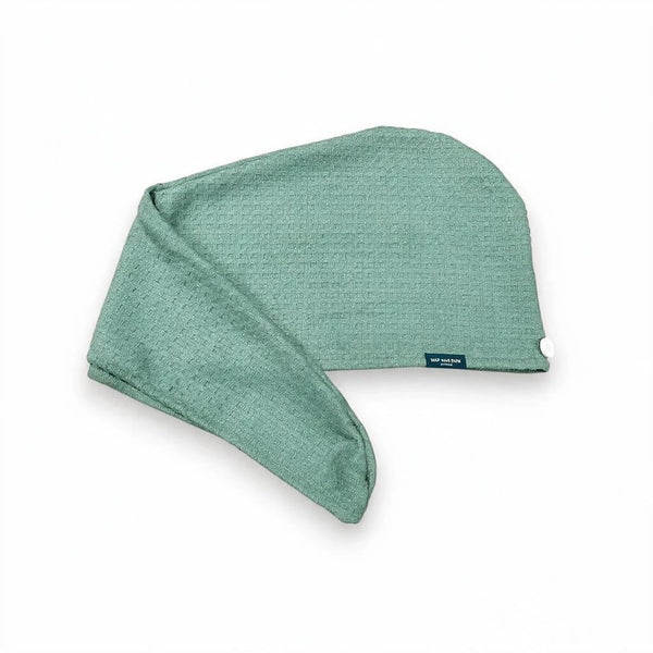 A sage green waffle hair towel wrap with a small dark blue brand tag, shown on a clean white background.