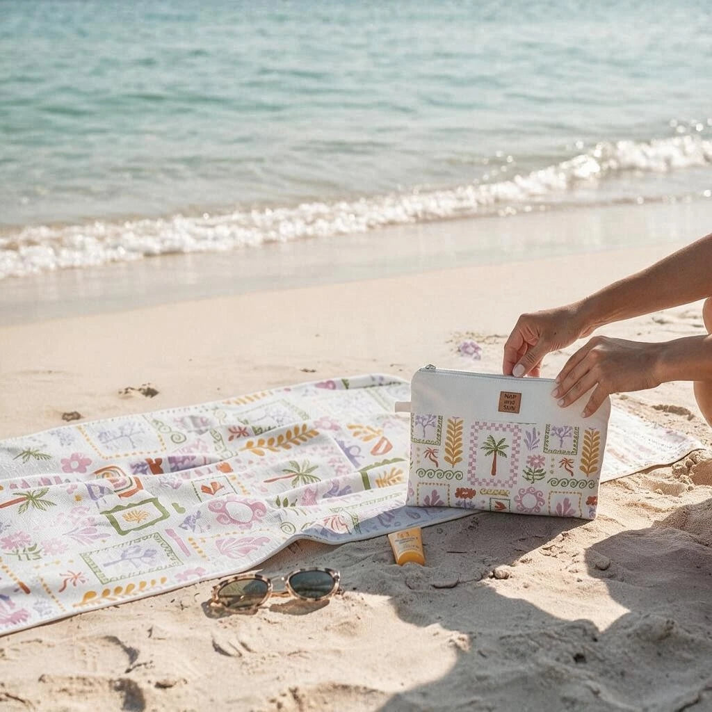 A person's hands are shown placing a white zippered pouch with a colorful tropical patchwork print onto a matching beach towel spread out on a sandy beach. The background features the blue ocean and shoreline.