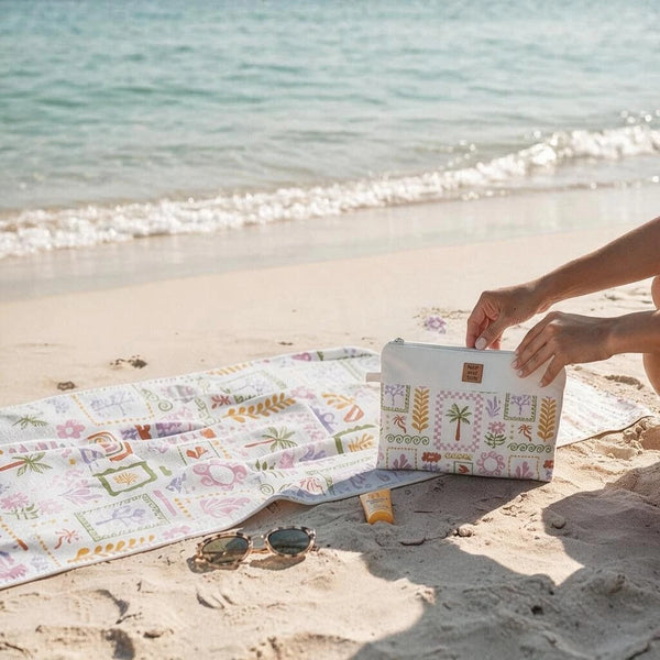 A person's hands are shown placing a white zippered pouch with a colorful tropical patchwork print onto a matching beach towel spread out on a sandy beach. The background features the blue ocean and shoreline.