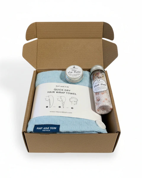 Open cardboard box containing a blue hair wrap towel, bath salt, lip balm on a white background