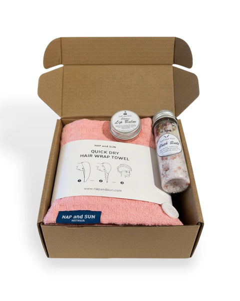 Open cardboard box containing a pink hair wrap towel, bath salt, lip balm on a white background