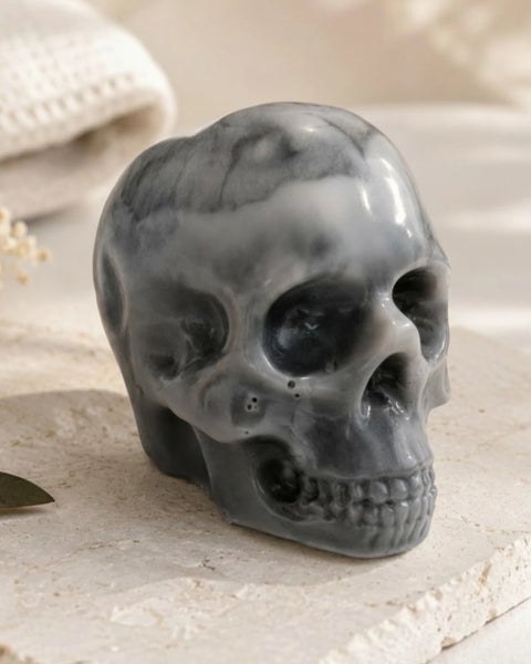A glossy dark gray skull-shaped decorative soap with marbled lighter gray veining, displayed on a light stone surface with soft neutral props in the background.