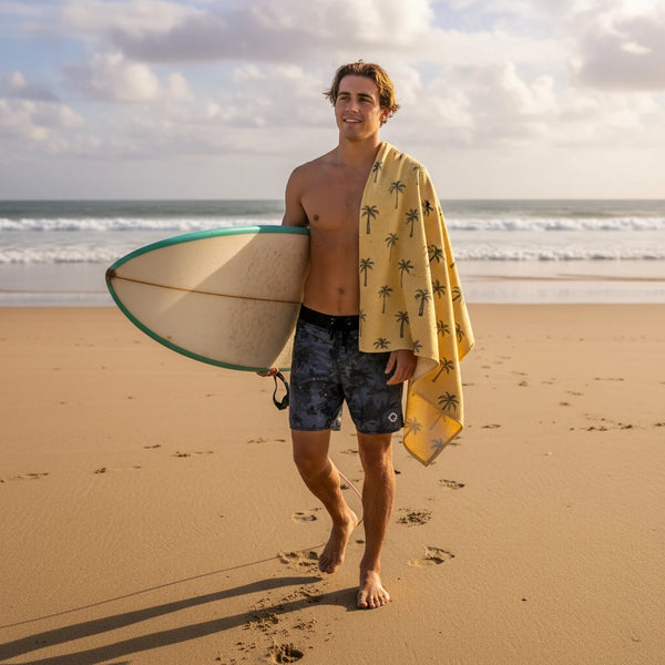 Man on a beach holding a surfboard with a towel draped over his shoulder