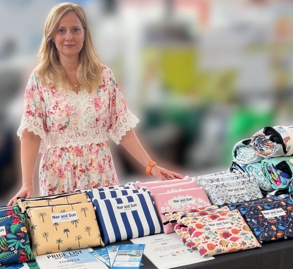 Andrea from Nap and Sun wearing a floral dress behind a market stall table at a Brisbane market, displaying Nap and Sun eco-friendly products such as sand-free beach towels and recycled waterproof picnic blankets with umbrella hole.