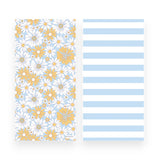 A beach towel  front and back sides shown side by side on a white background: the left has a floral print with white daisies and yellow flowers on a light blue ground, and the right has wide light blue and white horizontal stripes.