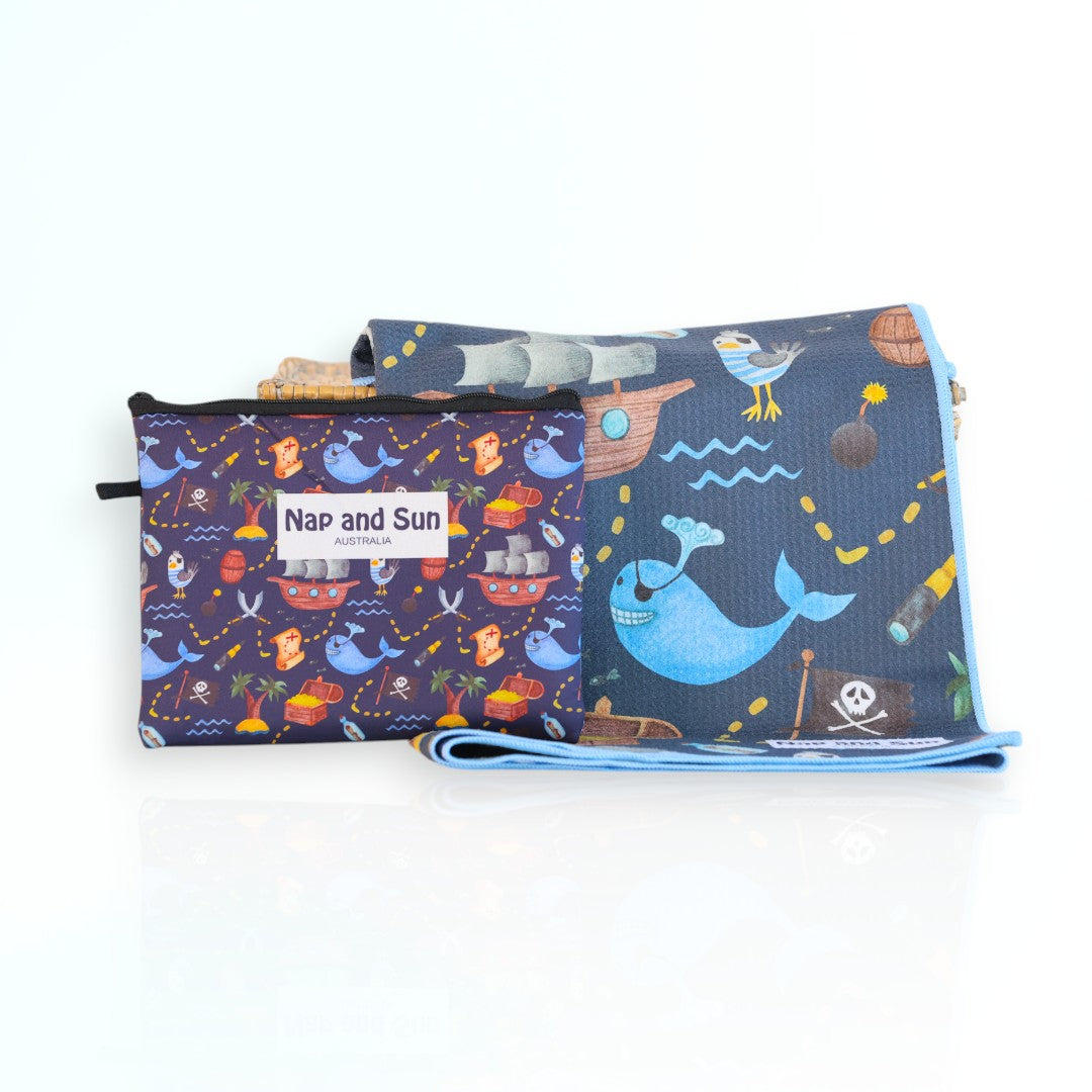 Napandsun Treasure Island sand-free beach towel for kids featuring a pirate-themed design with whales, islands, treasure map, and pirate ships on a dark blue background.  Matching wet bag is included. 