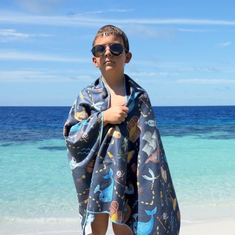 A child stands by a beach, wrapped snugly in a colorful pirate theme beach towel. Towel name: Napandsun Treasure Island sand-free beach towel for kids.