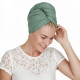 A woman wearing a textured sage green waffle towel turban, styled with a white lace top against a solid white background.