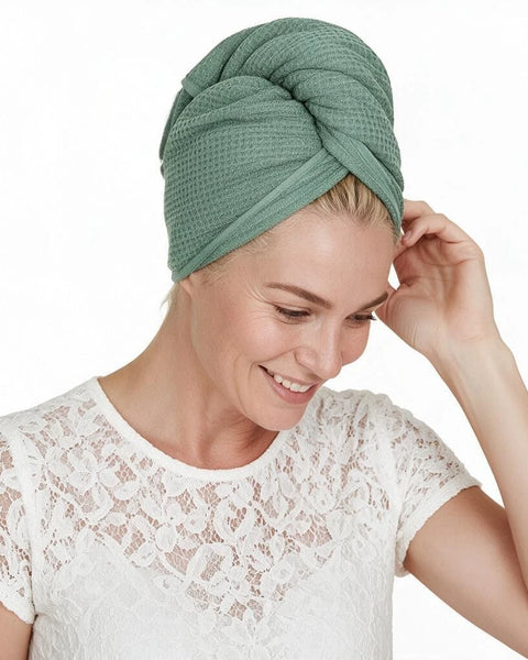 A woman wearing a textured sage green waffle towel turban, styled with a white lace top against a solid white background.